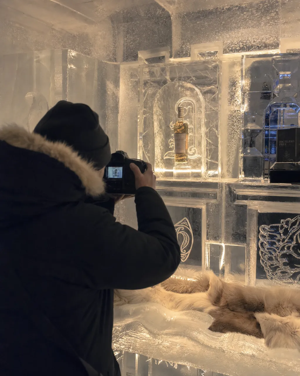 Interior of a premium branded ice bar, photographer seen from behind framing an illuminated product display carved into the ice, moody lighting and warm highlights on the ice textures