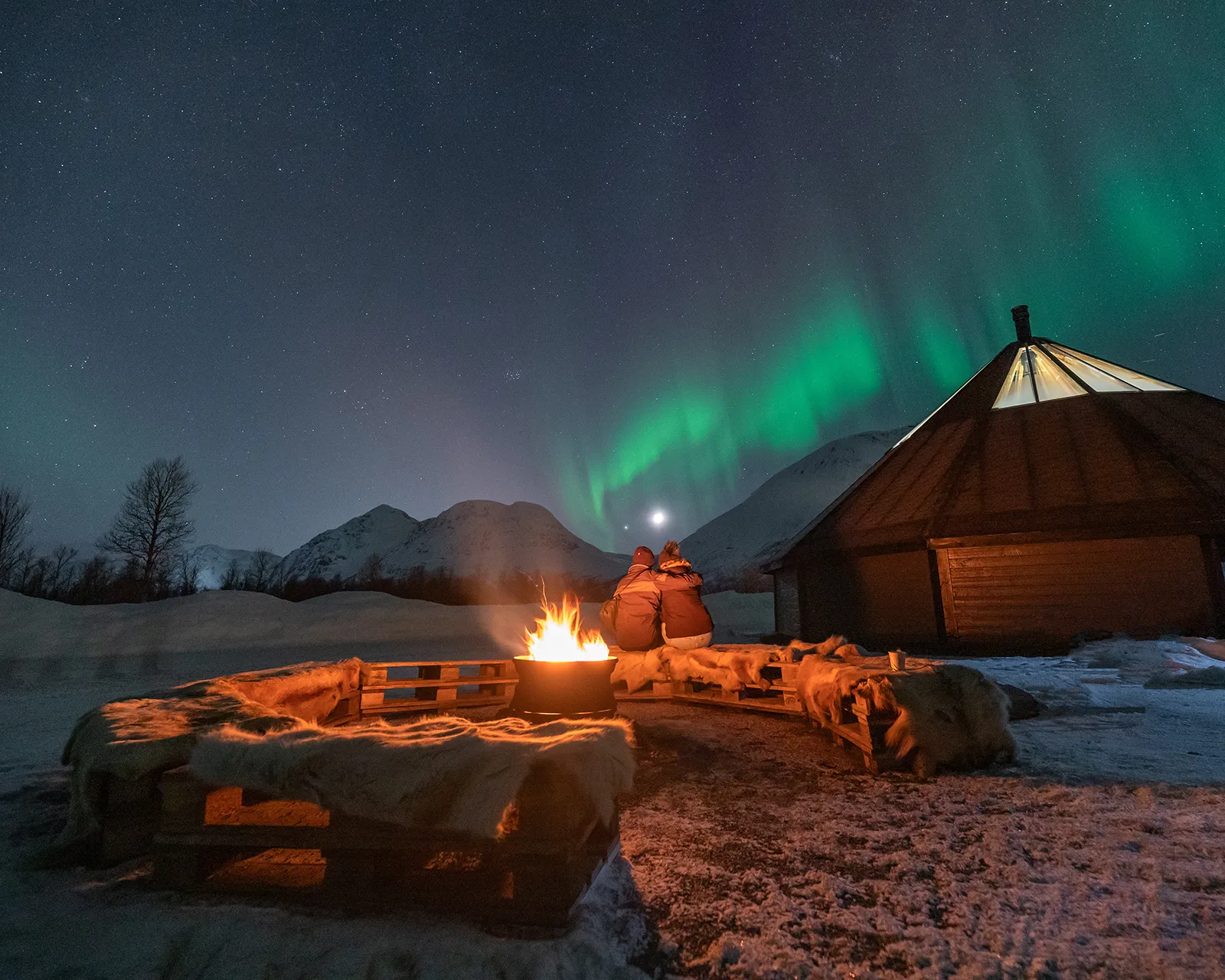 Two people sit shoulder to shoulder on reindeer hides around an open fire outside a lit Arctic hut, watching green northern lights over snow-covered mountains