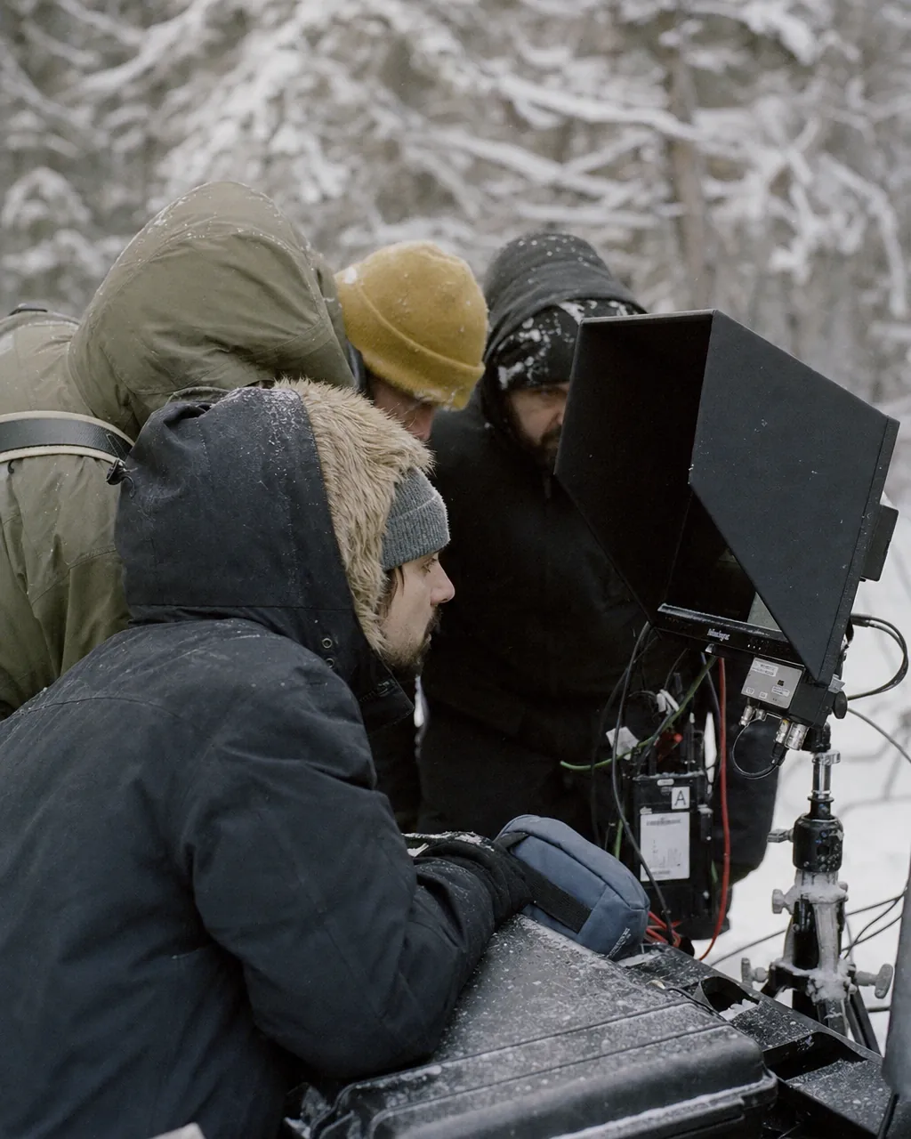 Film crew in weathered Arctic gear seen from the side, huddled around a high-brightness field monitor on a tripod in a snowy pine forest, focused on the frame