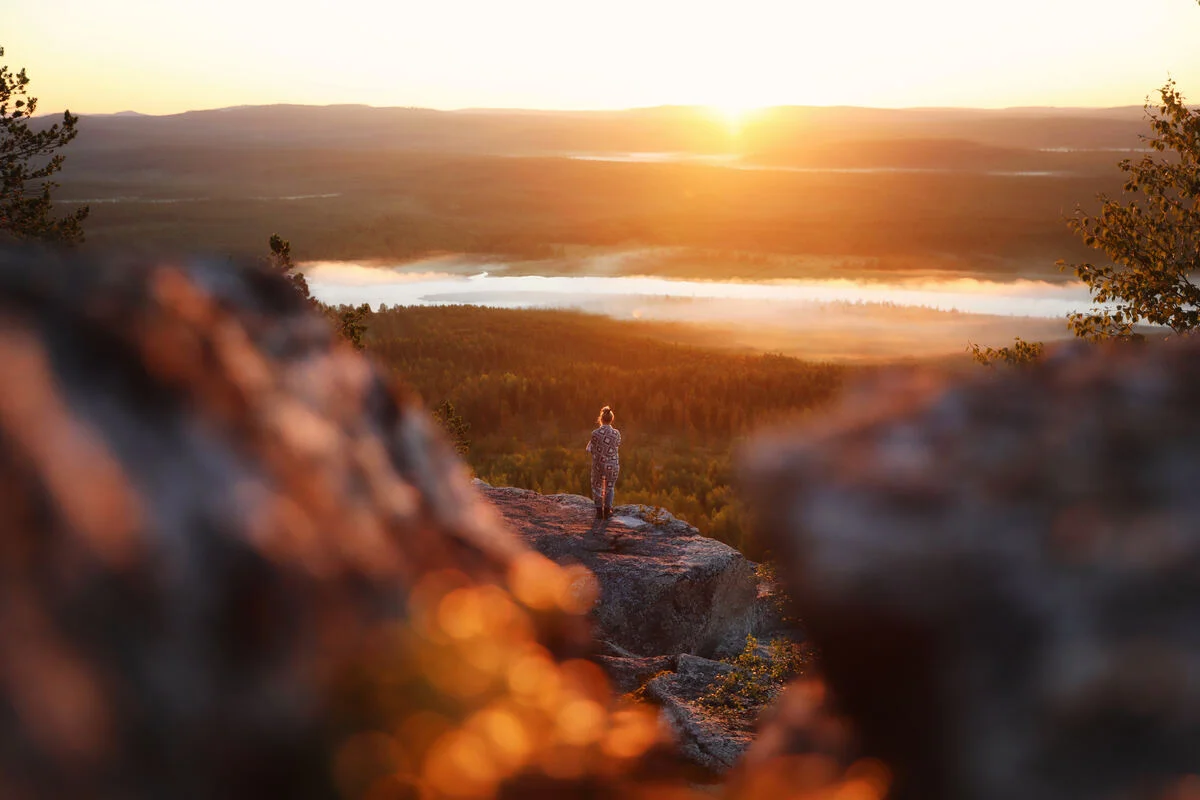 Single figure wrapped in a wool blanket on the Aavasaksa fell top at golden midnight sun, mist rising off a thawing river valley below, Finnish Lapland spring
